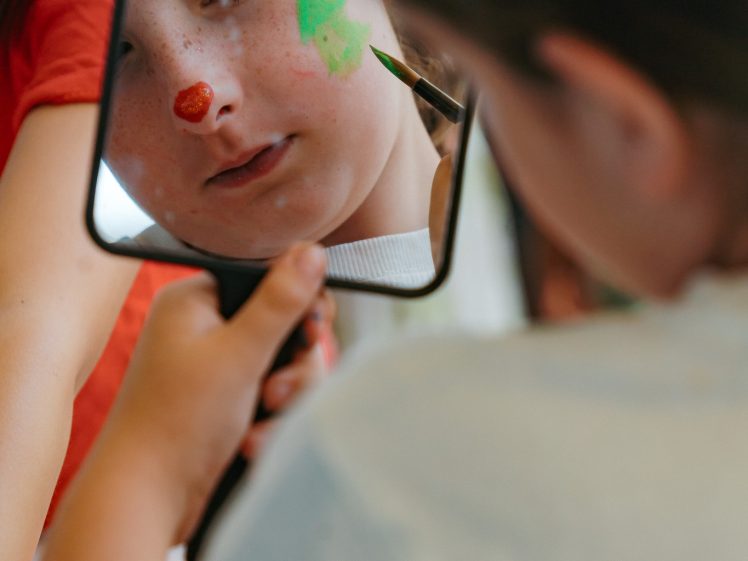 Image of A child looks into a handheld mirror while having their face painted. A small red dot is painted on their nose, and a green tree shape is painted on their cheek.