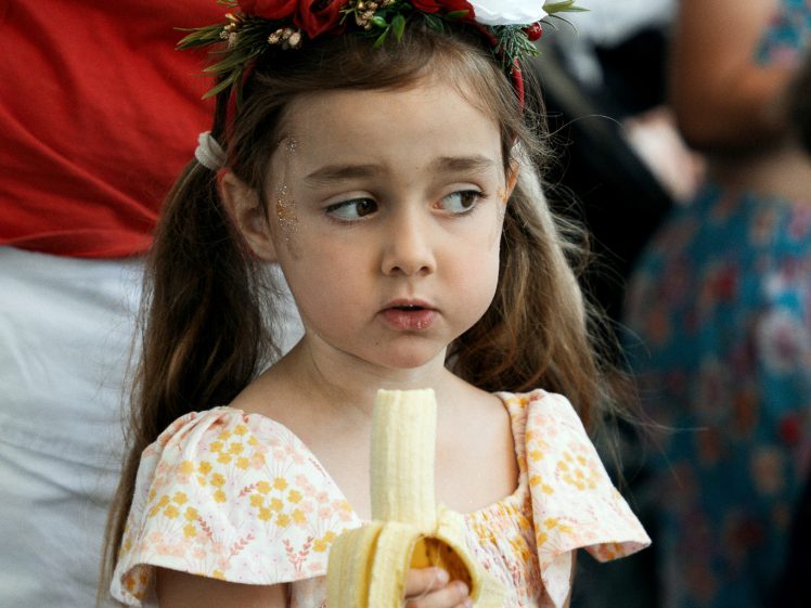 Image of A young girl with brown hair in plaits, wearing a floral dress and a flower crown, holds a peeled banana and looks to the side. An adult in a red shirt stands behind her.