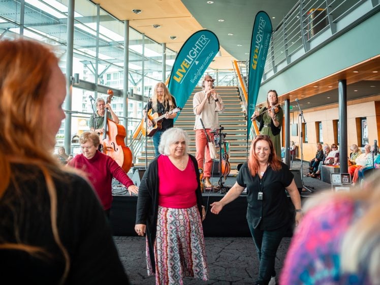 Image of A group of older adults dance in front of a live band performing indoors on a stage. Two vertical banners read Livelighter. People are enjoying the lively atmosphere in a modern, spacious venue.