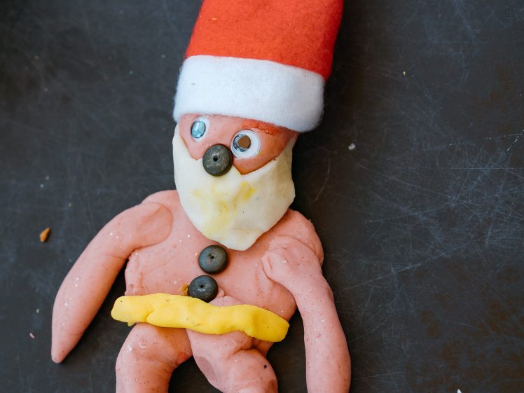 Image of A Father Christmas figure made from playdough and other craft materials, with a red hat, white beard, button eyes, and yellow belt, lying on a dark surface.