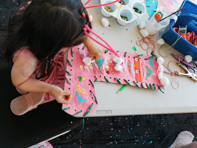 Image of A young girl decorates a large pink paper stocking with cotton wool, stickers, and glitter at a craft table, surrounded by art supplies like scissors, felt-tip pens, and small containers.