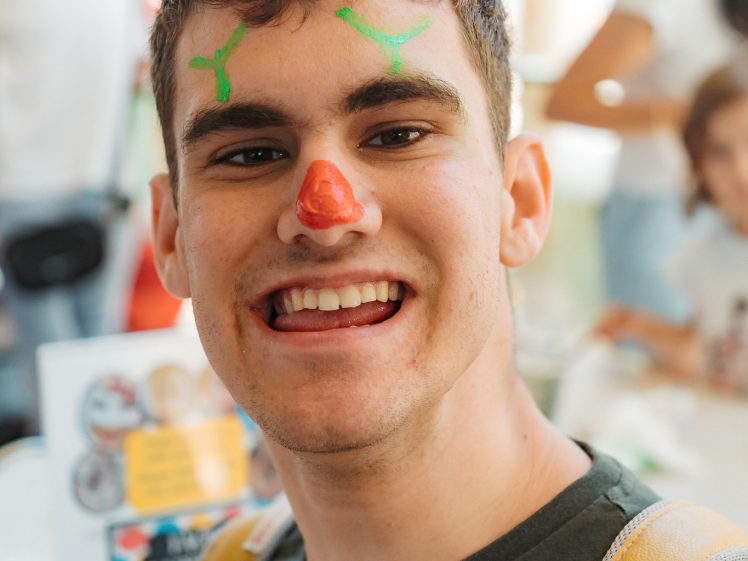 Image of A young person with face paint—a red nose and green marks on their forehead—smiles at the camera. They are wearing a dark shirt and rucksack, with a busy indoor setting in the background.