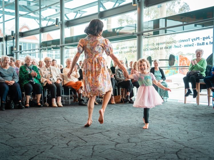 Image of A woman and a young girl in colourful dresses dance barefoot in front of an audience of older adults seated indoors by large windows. The child smiles whilst holding the woman's hand.