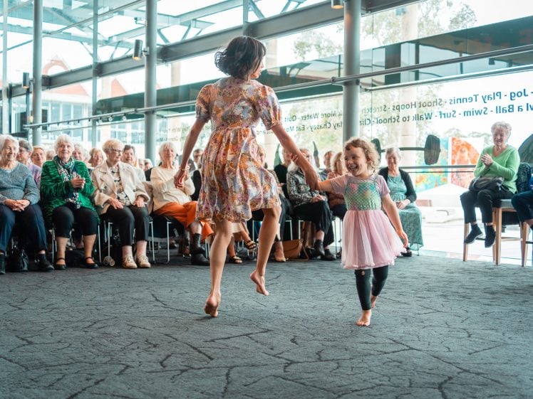 Image of A woman and a young girl joyfully dance hand in hand in front of an audience of elderly people, who are seated and watching them in a bright, modern room with large windows.