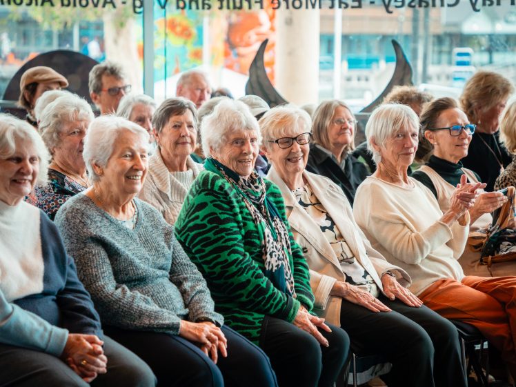 Image of A group of elderly women sit closely together in rows, smiling and applauding, as they attend an indoor event with large windows in the background.