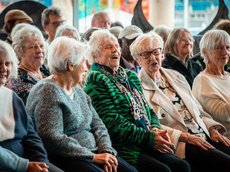 Image of A group of elderly women sit closely together in an audience, smiling and laughing, enjoying a lively event. Other people are visible in the background, adding to the cheerful atmosphere.