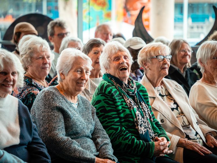 Image of A group of elderly women sit closely together, smiling and laughing, in a bright indoor setting with large windows and colourful decor in the background.