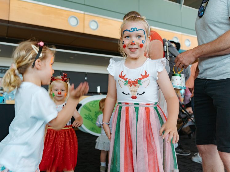 Image of Three young girls wearing face paint and festive outfits stand indoors, with adults nearby. One girl at the front wears a colourful striped dress and smiles at the camera. The setting appears to be a cheerful event or party.