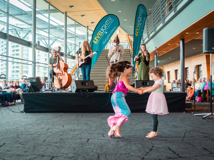 Image of Two young girls dance together barefoot in front of a stage where a band performs live music. The scene takes place indoors with an audience seated nearby and large windows in the background.