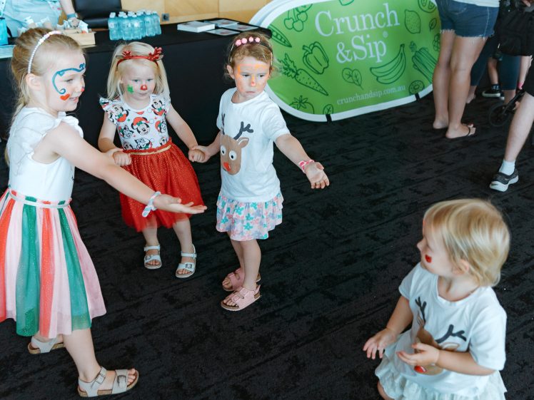 Image of Four young children with face paint and festive clothes stand and kneel in a circle, playing together indoors near a green Crunch & Sip banner and a table with water bottles.