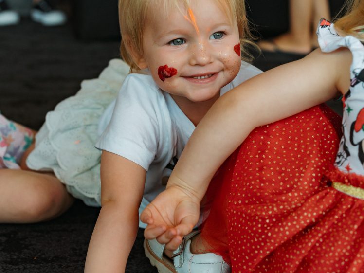 Image of A smiling young child with blonde hair, heart stickers on their cheeks, and a painted orange line on their forehead is lying on the floor next to another child in a red dress.