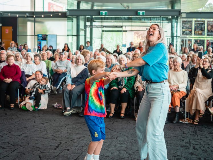 Image of A young boy in a tie-dye shirt and a woman in light blue jeans joyfully dance together in front of a seated audience of mostly older adults, all watching and smiling in a brightly lit indoor space.