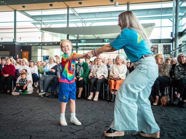 Image of A young boy and a woman, both wearing colourful clothes, joyfully dance together in front of an audience seated in a bright, modern room with large windows. The boy is smiling and holding the woman’s hand.