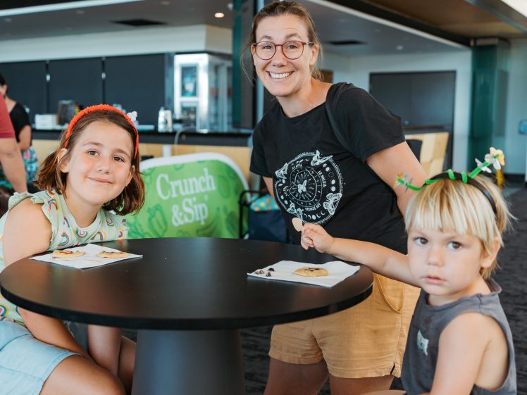 Image of A smiling adult and two young children sit at a round table with snacks indoors. A green Crunch & Sip banner is in the background. The atmosphere appears casual and friendly.
