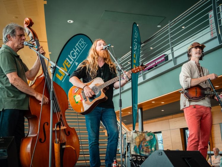 Image of Three musicians perform indoors; one plays a double bass, another sings and plays acoustic guitar, and the third plays a violin. Banners and people seated in the background are visible.