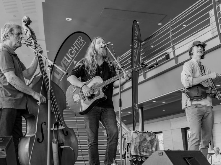 Image of Three musicians perform indoors; one plays a double bass, another sings and plays acoustic guitar, and the third plays the violin. Banners and stairs are visible in the modern background. The image is in black and white.