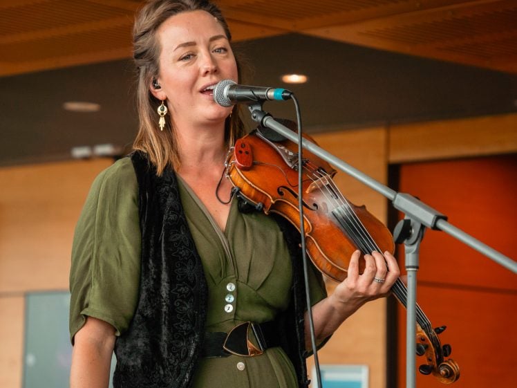 Image of A woman wearing a green dress and black waistcoat sings into a microphone while holding a violin on stage. She wears earrings and has light brown hair, with warm indoor lighting in the background.
