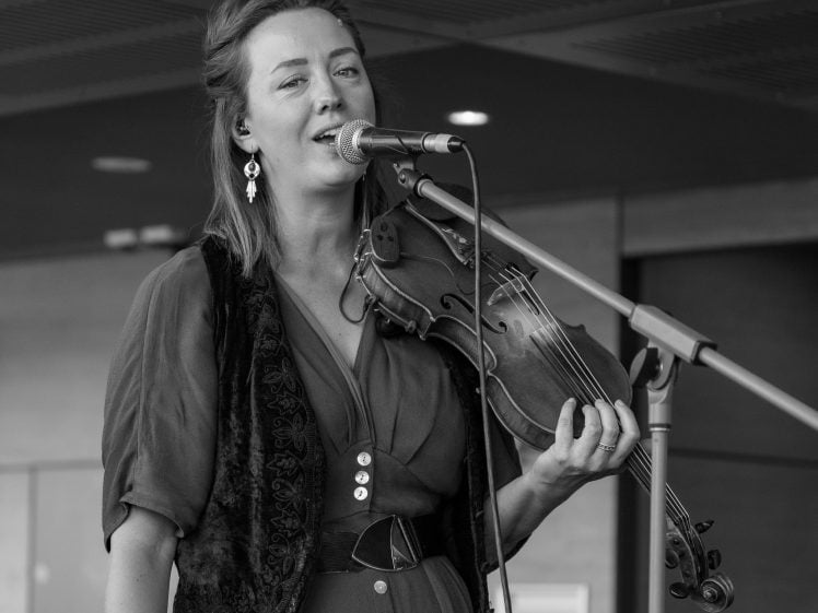Image of A woman stands singing into a microphone while holding a violin. She wears a patterned waistcoat, a dress with buttons, and earrings. The image is in black and white and captures a performance setting.