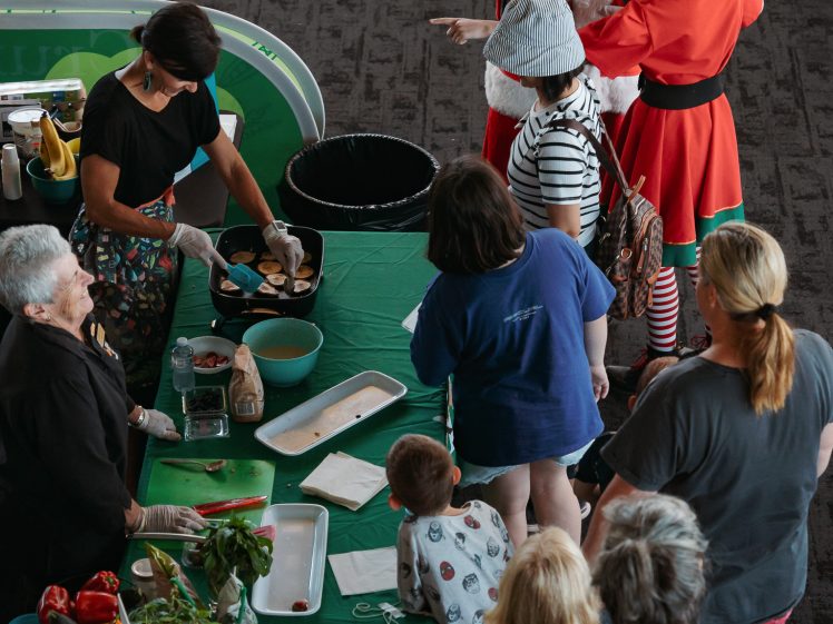 Image of People gather around a green table with food, while Father Christmas and an elf greet children nearby. Some people are serving food, and others are queueing. The scene takes place indoors with festive holiday decor.