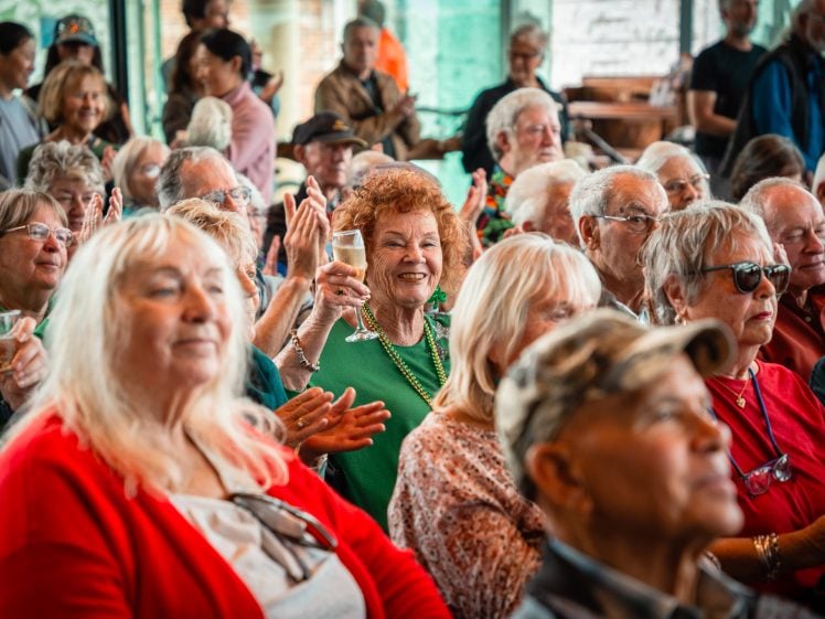 Image of A group of elderly people sit closely together at an indoor event. One woman in the centre, wearing green, smiles and raises a glass in celebration. Others around her clap, chat, and look towards the front.