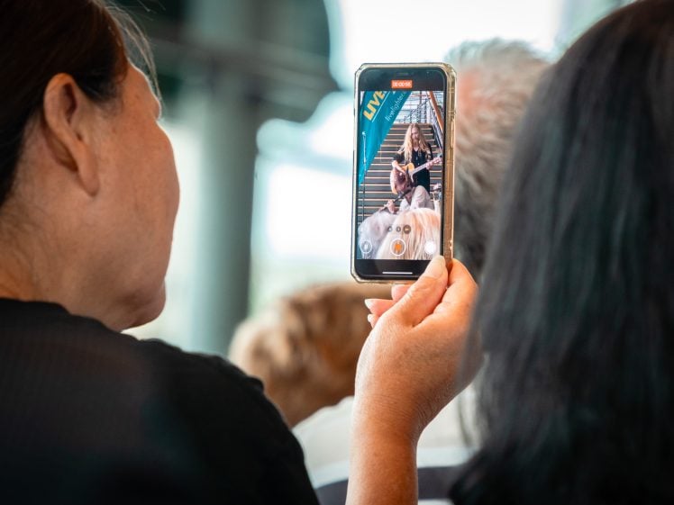 Image of A person holds up a mobile phone, recording or taking a photo of a woman playing guitar on a staircase. The phone screen clearly displays the woman, whilst people are visible in the foreground.
