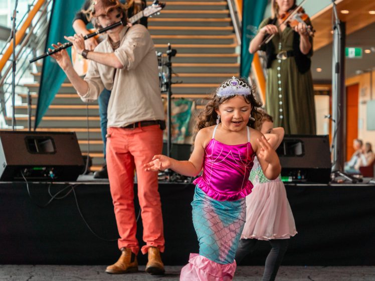 Image of A young girl wearing a mermaid costume and tiara dances in front of a stage where musicians play flute and violin. The background shows stairs and musical equipment.