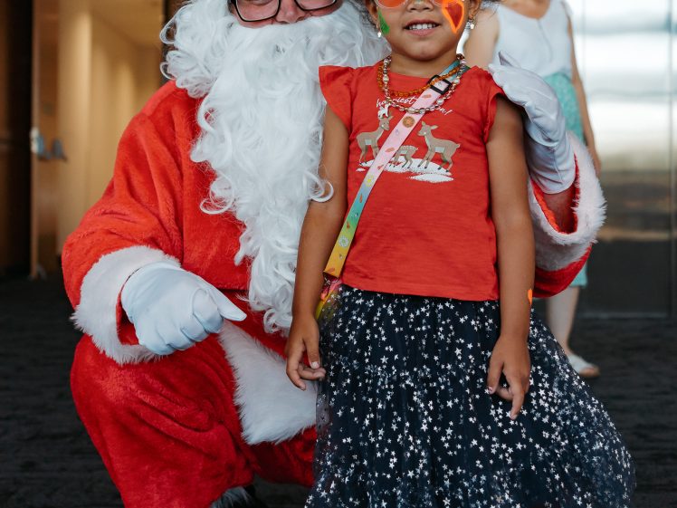 Image of A person dressed as Father Christmas kneels beside a smiling young girl in festive clothing and colourful sunglasses. Another woman stands in the background near a lift.