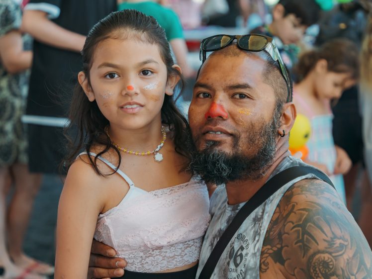 Image of A man with tattoos and sunglasses on his head hugs a young girl. Both have painted noses and glitter on their cheeks. People and children are visible in the background, suggesting a festive event.
