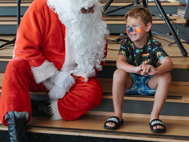 Image of A person dressed as Father Christmas sits on wooden steps next to a young boy with face paint, wearing shorts and sandals. They appear to be having a friendly conversation indoors.