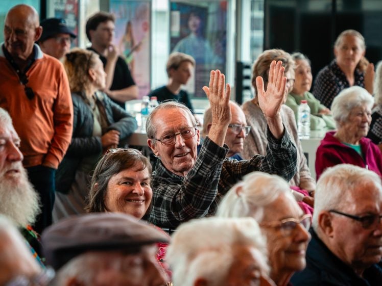 Image of A group of elderly people sitting closely together indoors, many smiling. One man in the centre, wearing glasses and a checked shirt, is enthusiastically clapping his hands above his head.