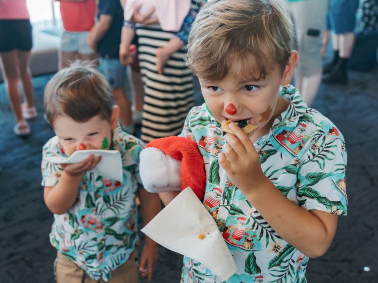 Image of Two young boys wearing matching tropical shirts eat snacks at an indoor event. The older boy holds a serviette and a Father Christmas hat, both have red noses painted on. Other people, mostly children, are visible in the background.