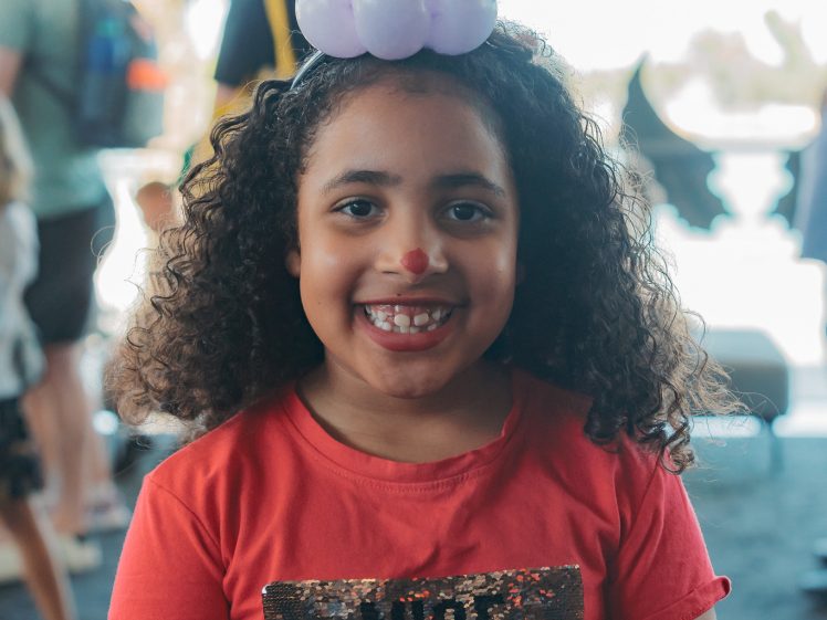 Image of A smiling child with curly hair wears a red shirt with the word NICE and a heart-shaped purple balloon hat featuring a cat face. The child has a red nose drawn on and people are visible in the blurred background.