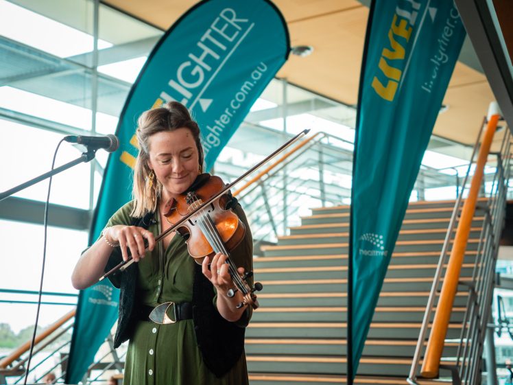 Image of A woman in a green dress plays a violin near a microphone, standing indoors by a staircase. Behind her are two large blue banners with “LIFELIGHTER” and “LIFEFLIGHT” written on them.