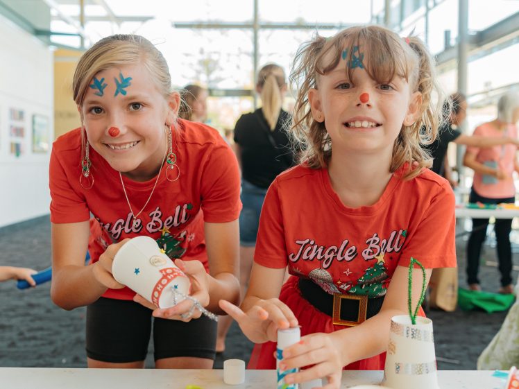 Image of Two children with festive face paint and red Jingle Bells shirts smile whilst making crafts with white cups and glue at a brightly lit indoor event. Other people are visible in the background.