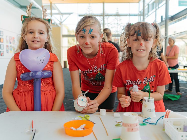 Image of Three young girls in red holiday tops stand at a craft table, smiling. One has reindeer antlers, another has blue face paint, and the third holds a balloon animal. Craft supplies are scattered on the table.
