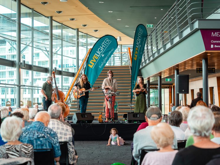 Image of A band performs onstage indoors in front of seated audience members. Large windows let in natural light. Two “LiveLighter” banners are behind the band, and a child sits on the floor near the stage, facing the crowd.