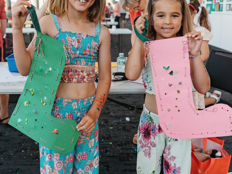 Image of Two young girls smile and hold up large, decorated paper boots—one green, one pink—at an arts and crafts event. Both are wearing colourful floral outfits and standing indoors, with craft supplies and confetti around them.