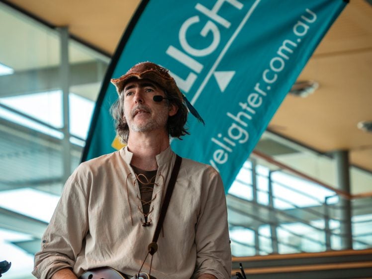 Image of A man wearing a brown hat and beige shirt plays a mandolin in front of a blue ELIGHTER banner and a staircase indoors.