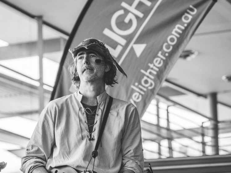 Image of A man playing a mandolin stands in front of a large flag reading elighter.com.au near a staircase in an indoor setting. He wears a hat and casual shirt. The photo is in black and white.