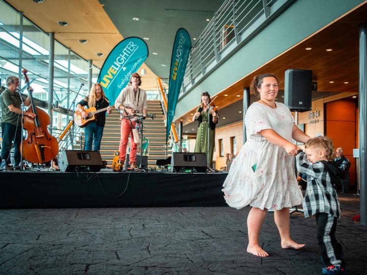 Image of A woman in a white dress dances barefoot with a small child in checked. Behind them, a band performs on stage with guitars, a double bass, and a violin. The scene is indoors with banners and a modern interior.