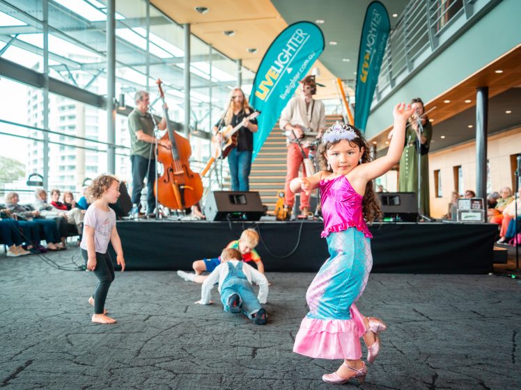 Image of Children dance and play near a stage where a live band performs in a bright, modern indoor venue. One child in a mermaid costume poses energetically at the front, whilst an audience watches from seated rows.