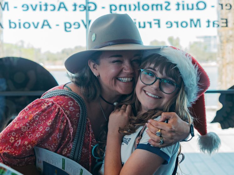 Image of Two women smile and hug indoors. One wears a hat and patterned top, holding a map; the other wears glasses, a Santa hat, and a white shirt. Sunlight comes through a window with text in the background.