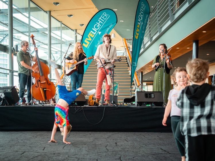 Image of A band performs on an indoor stage with instruments whilst children play and dance in front, including one child doing a handstand. “LiveLighter” banners are displayed behind the musicians. The setting appears bright and modern.