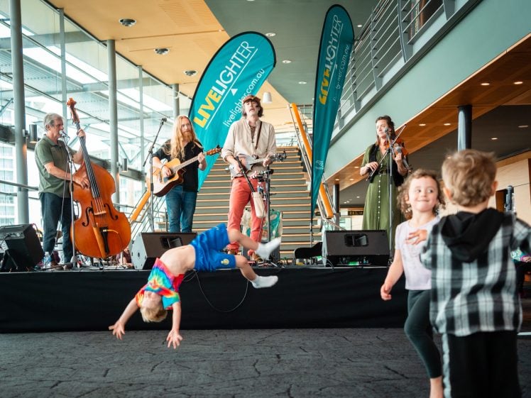 Image of A lively band performs on stage indoors as young children dance and play in front; one child in a tie-dye shirt is doing a cartwheel whilst others smile and move around joyfully.