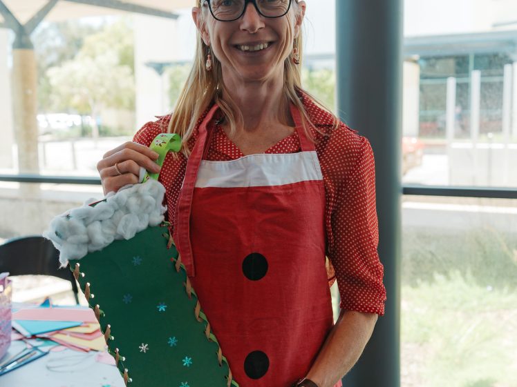 Image of A smiling woman wearing reindeer antlers and a red apron with black buttons holds a large green Christmas stocking decorated with white trim and blue stars. She stands indoors near a table with craft supplies.