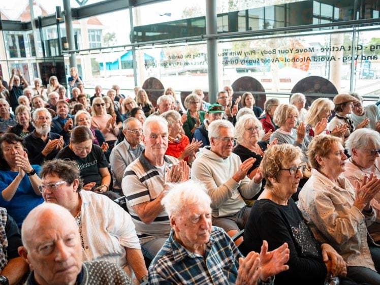 Image of A large group of older adults sit closely together indoors, many clapping and smiling, in what appears to be an audience at a public event. Bright natural light fills the room through large windows.