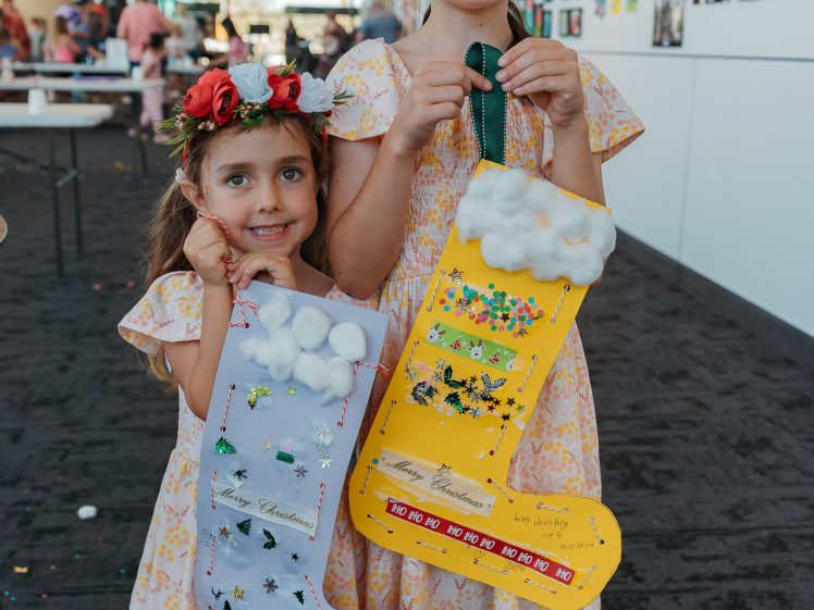 Image of Two young girls in matching yellow floral dresses and flower crowns smile and hold decorated Christmas stockings. One stocking is yellow with cotton at the top, the other is purple. Artworks are displayed on the white wall behind them.