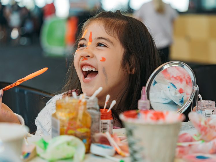 Image of A young girl with paint on her face laughs joyfully while painting. Art supplies, brushes, and a mirror are on the table in front of her, creating a lively and creative atmosphere.
