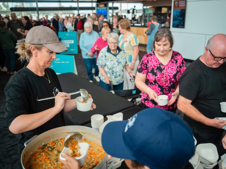 Image of A woman serves soup from a large pot into bowls for people standing in queue at an indoor event. Several people wait their turn, holding empty bowls, whilst others receive their servings.