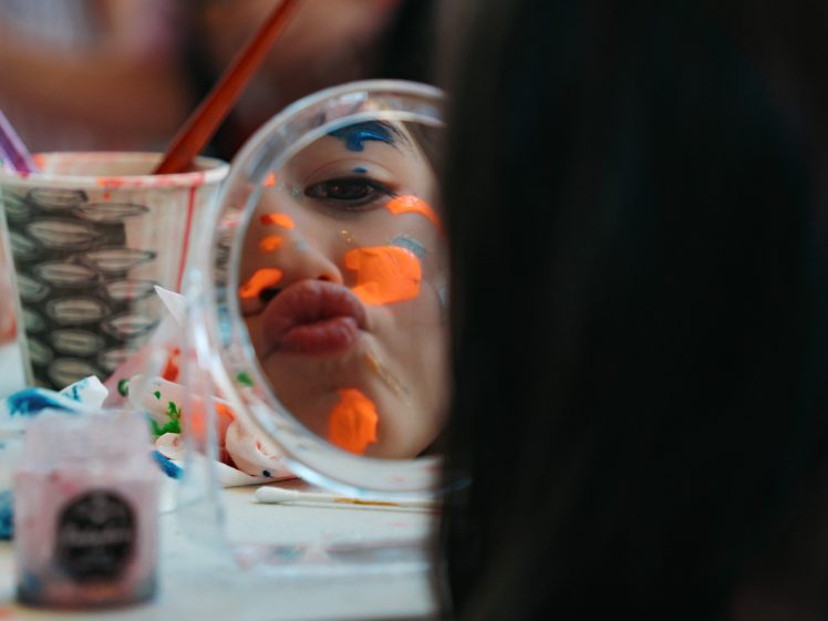 Image of A child with colourful paint on their face makes a pouty expression into a small round mirror, surrounded by art supplies and painted cups on a table.